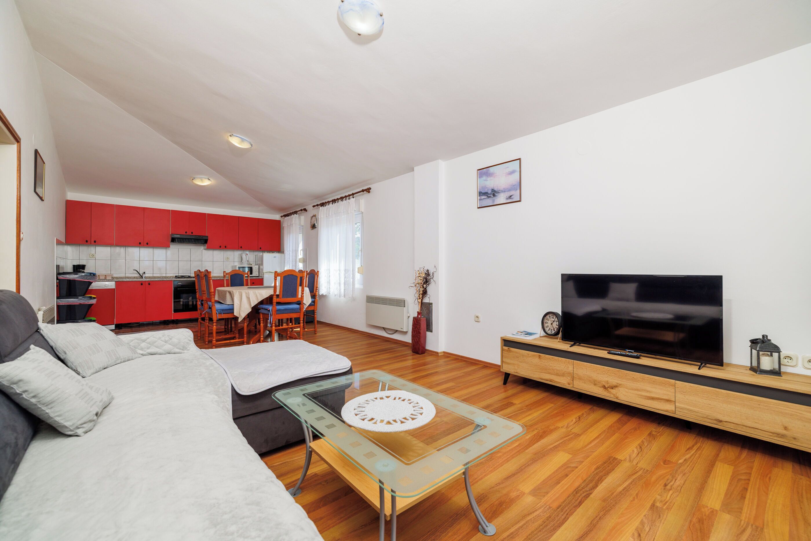 Een moderne woonkamer met een bank, een glazen salontafel, een televisie op een houten meubel en een keuken op de achtergrond met rode keukenkastjes.