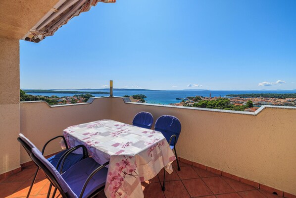 A balcony with a table and chairs, overlooking a coastal view with houses and the sea. - Apartments Marija (Mundanije)
