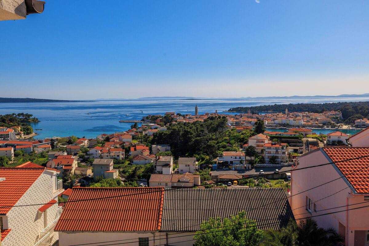 A coastal town with red-tiled roofs, a clear blue sky, and a view of the sea.