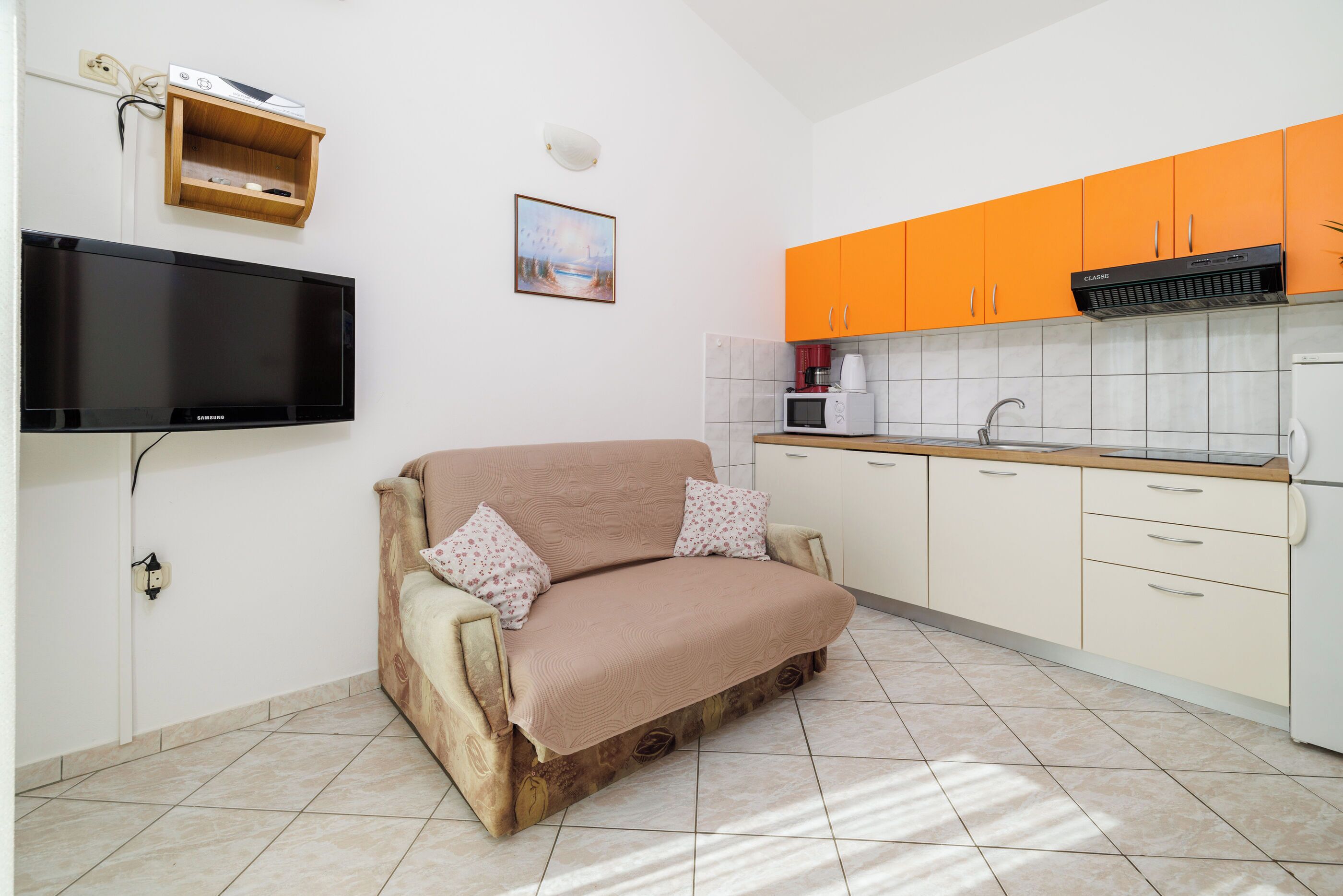 A compact kitchen with orange cabinets, a flat-screen TV, and a sofa with a blanket.