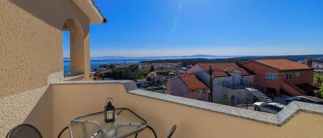 A balcony with a table and chairs, overlooking a coastal town with houses and a clear blue sky.