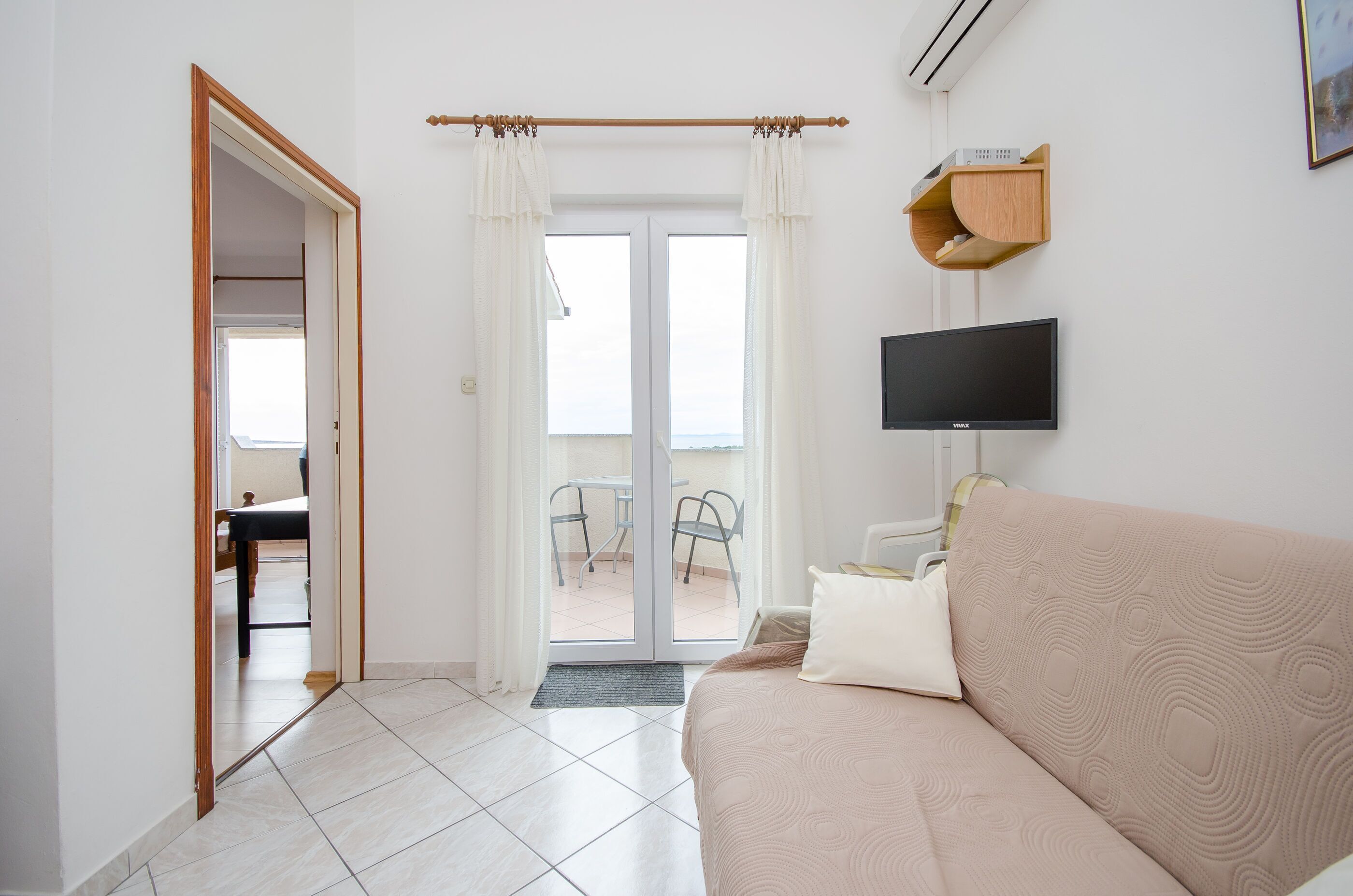 Una habitación de hotel moderna con sofá, televisor de pantalla plana, estante de madera y puerta de cristal corredera que da a un balcón.