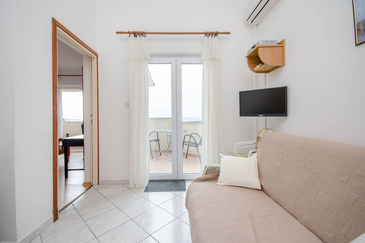 A modern hotel room with a sofa, a flat-screen TV, a wooden shelf, and a sliding glass door leading to a balcony.