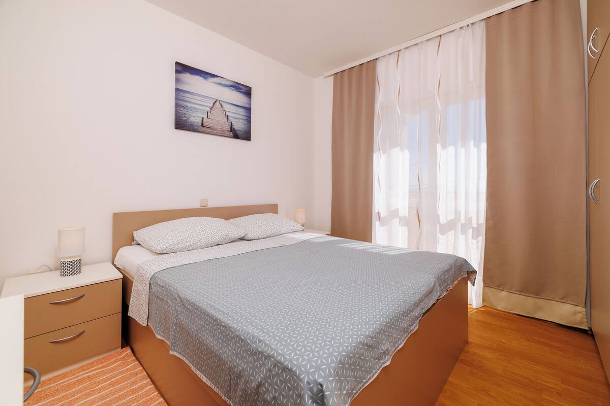 A bedroom with a bed, a nightstand, a drawer unit, a painting, and a window with curtains.