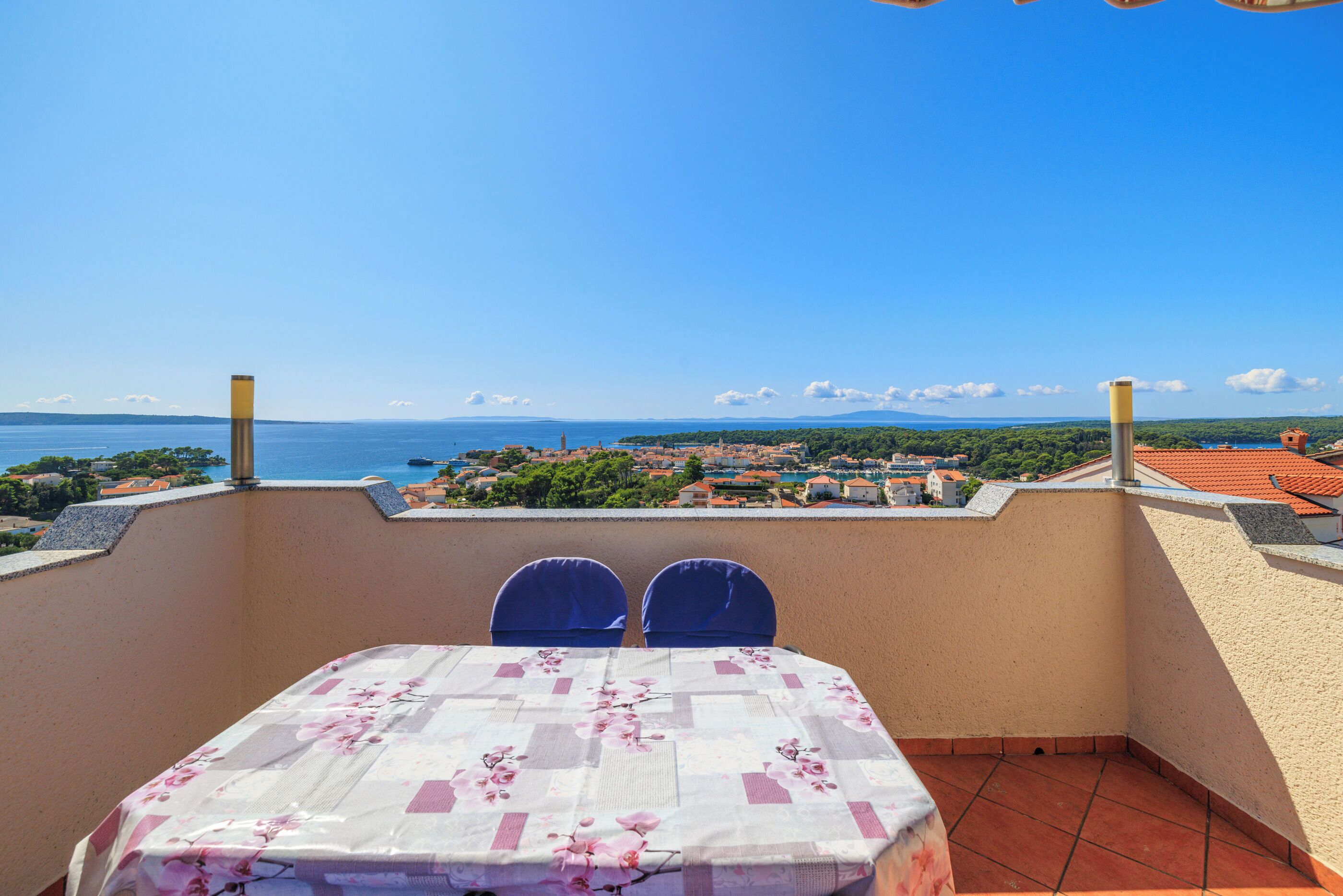 A balcony with a table and chairs, overlooking a coastal town and the sea.