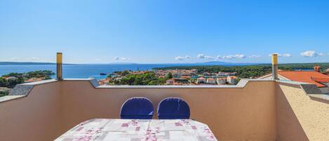 A balcony with a table and chairs, overlooking a coastal town and the sea.