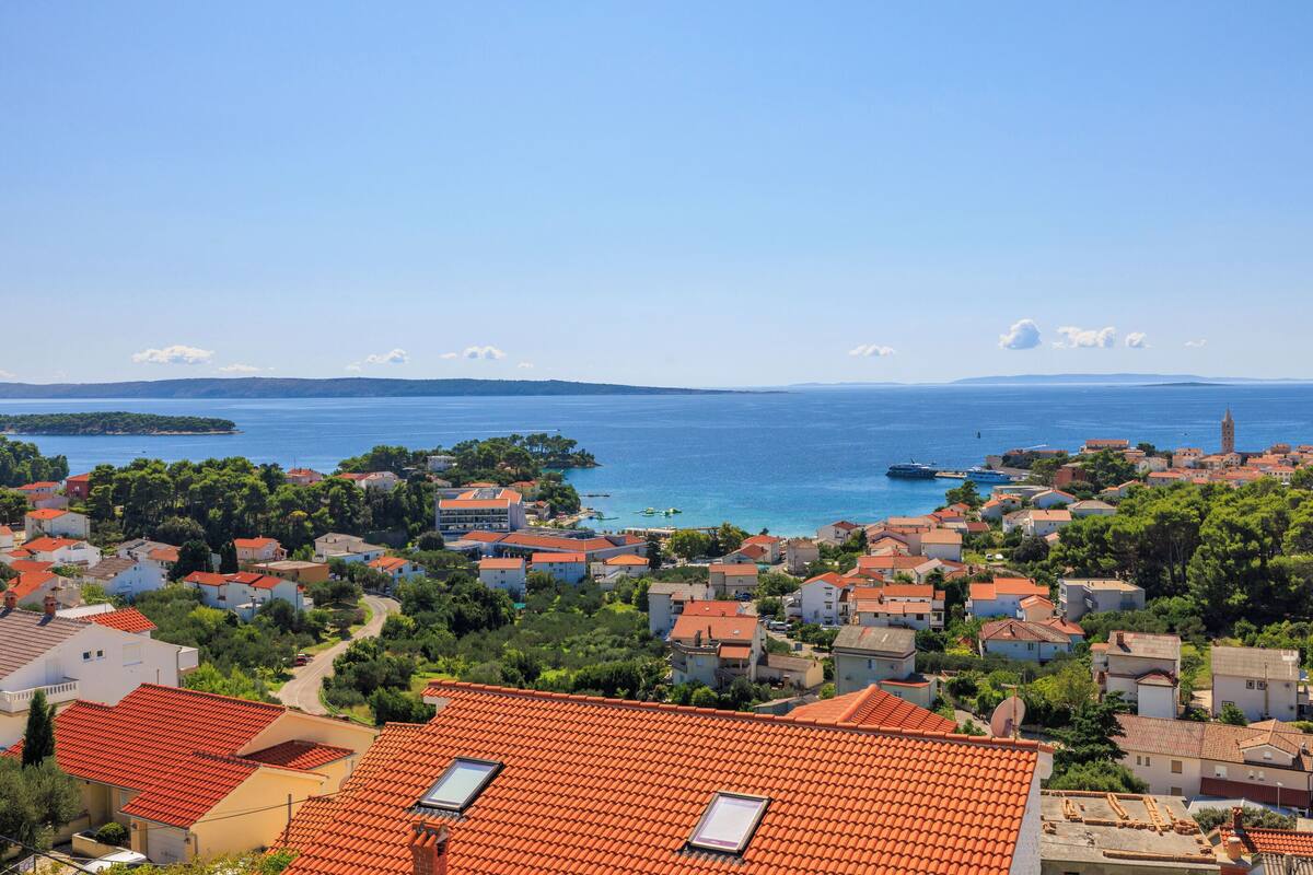 A coastal town with red-roofed buildings, a clear blue sea, and a distant island.