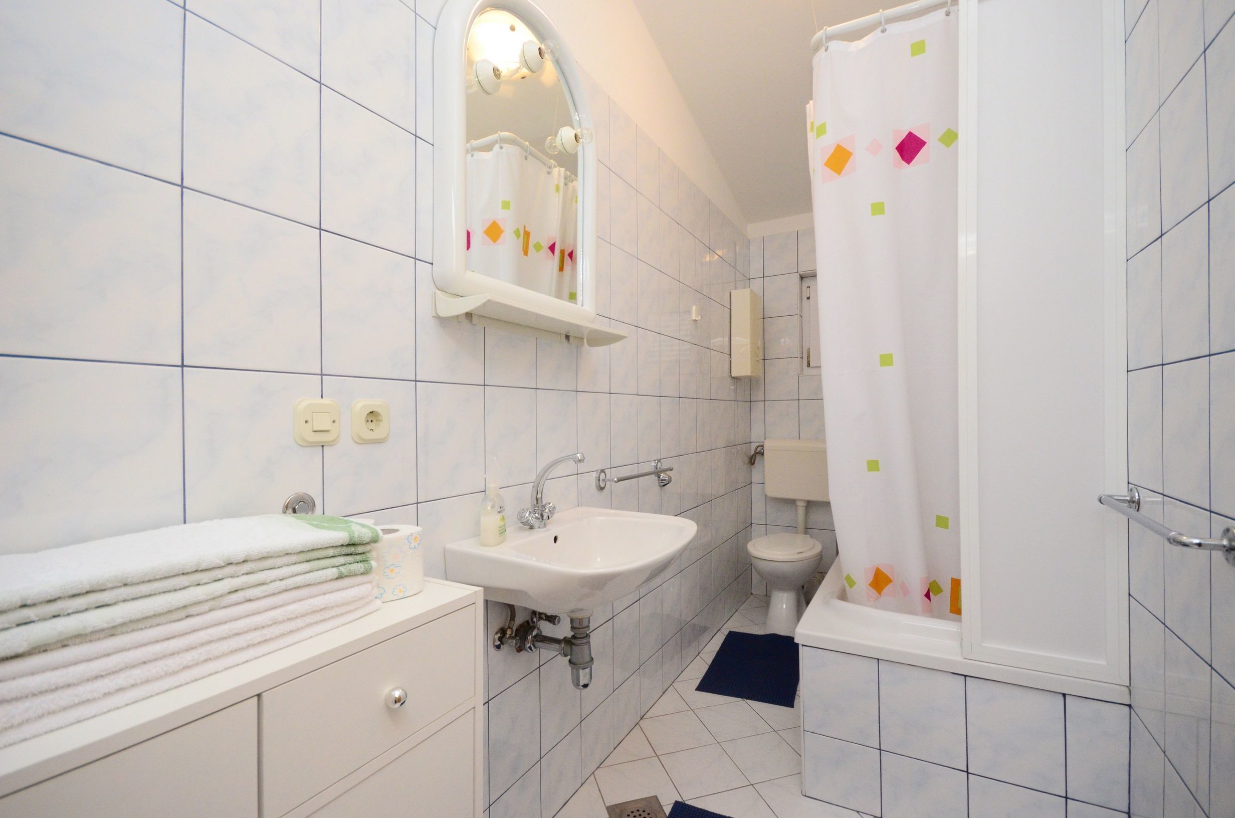 Un baño con azulejos blancos, un lavabo, un espejo, un inodoro y una cortina de ducha con diseños coloridos.