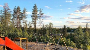 Une aire de jeux avec un toboggan rouge et des balançoires, entourée d’arbres et sous un ciel dégagé.