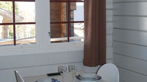 Un coin repas avec une table blanche dressée pour deux personnes, une fenêtre avec des stores et un luminaire suspendu.