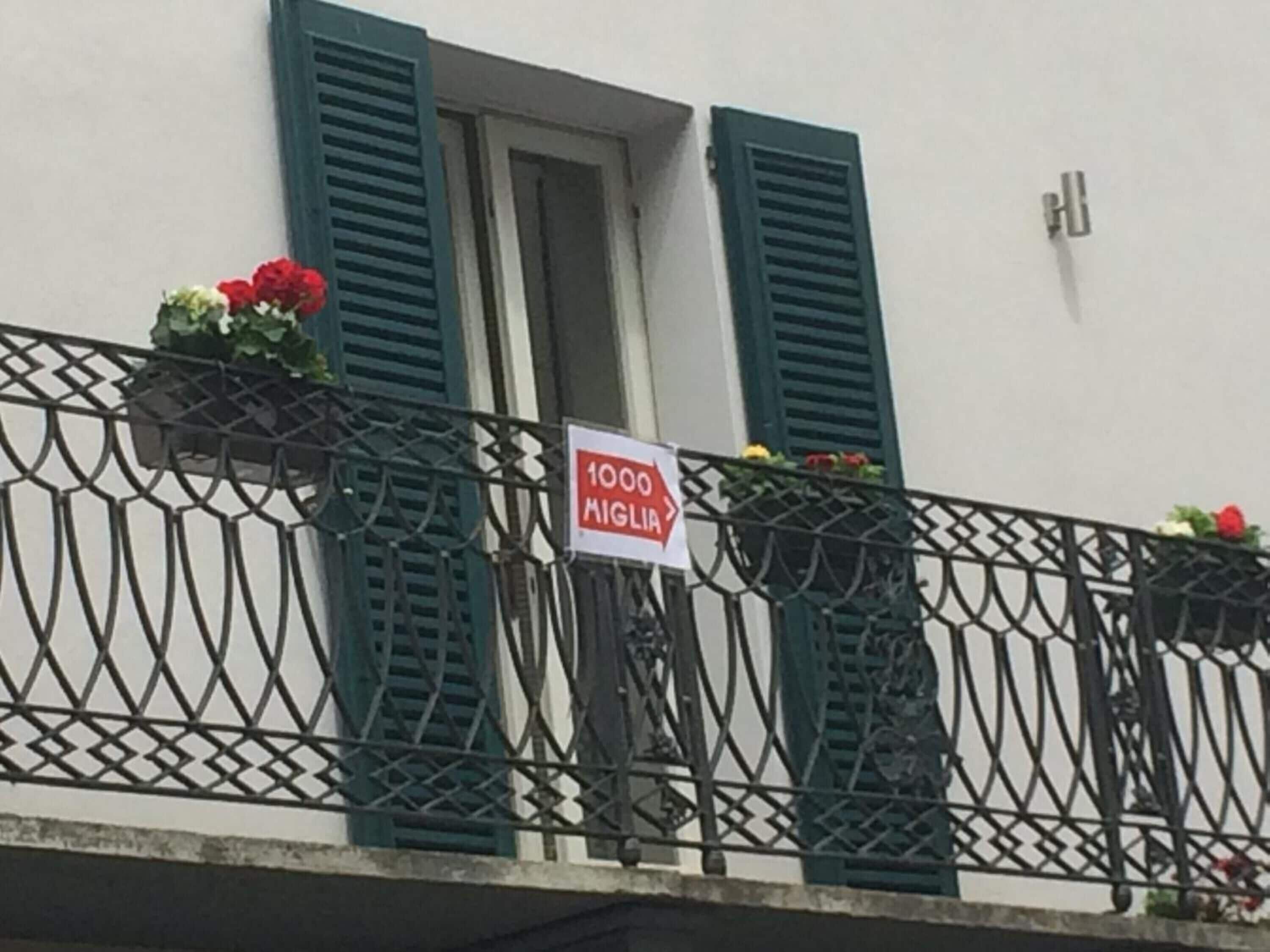 Ein Balkon mit schmiedeeisernem Geländer, grünen Fensterläden und Blumenkästen. Ein Schild trägt die Aufschrift "1000 Miglia".