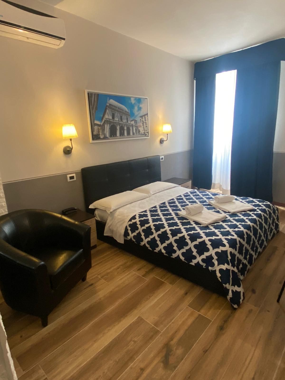 Una camera d'albergo con un letto, una sedia, un quadro appeso al muro e una finestra con tende.