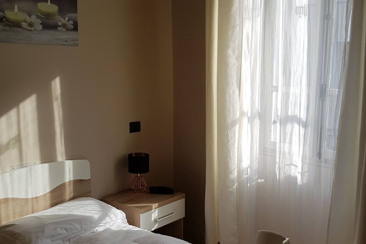 A bedroom with a bed, a nightstand, a lamp, and a window with sheer curtains.