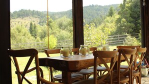 Una mesa de madera con seis sillas, vista a una ladera boscosa y una cerca de madera al fondo.