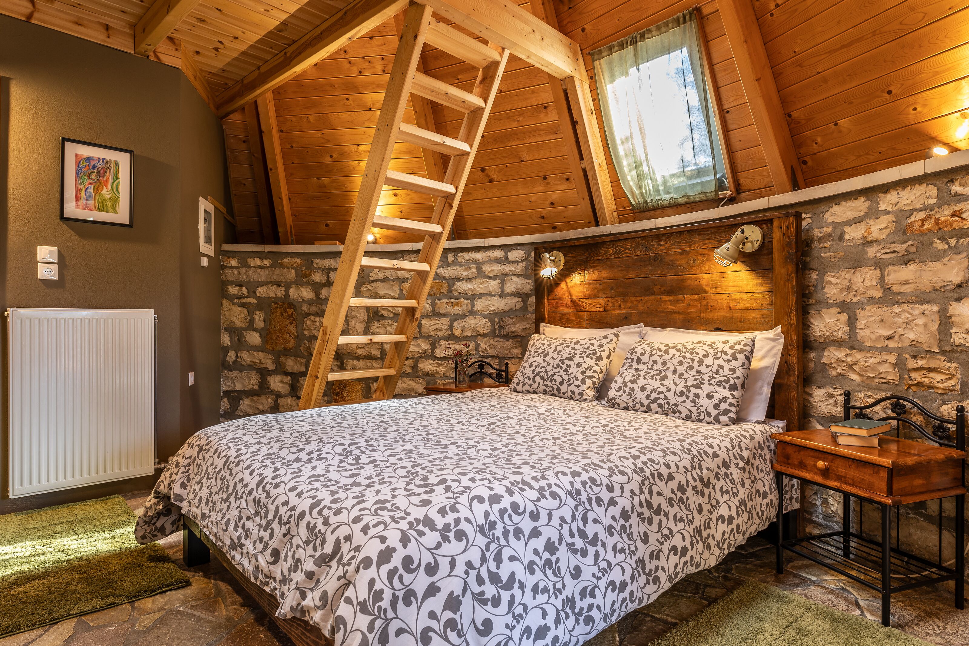 A bedroom with a wooden ladder, a stone wall, a bed with patterned bedding, a bedside table with a book, and a window with a curtain.