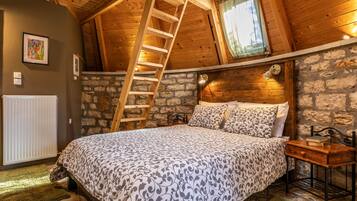 A bedroom with a wooden ladder, a stone wall, a bed with patterned bedding, a bedside table with a book, and a window with a curtain.