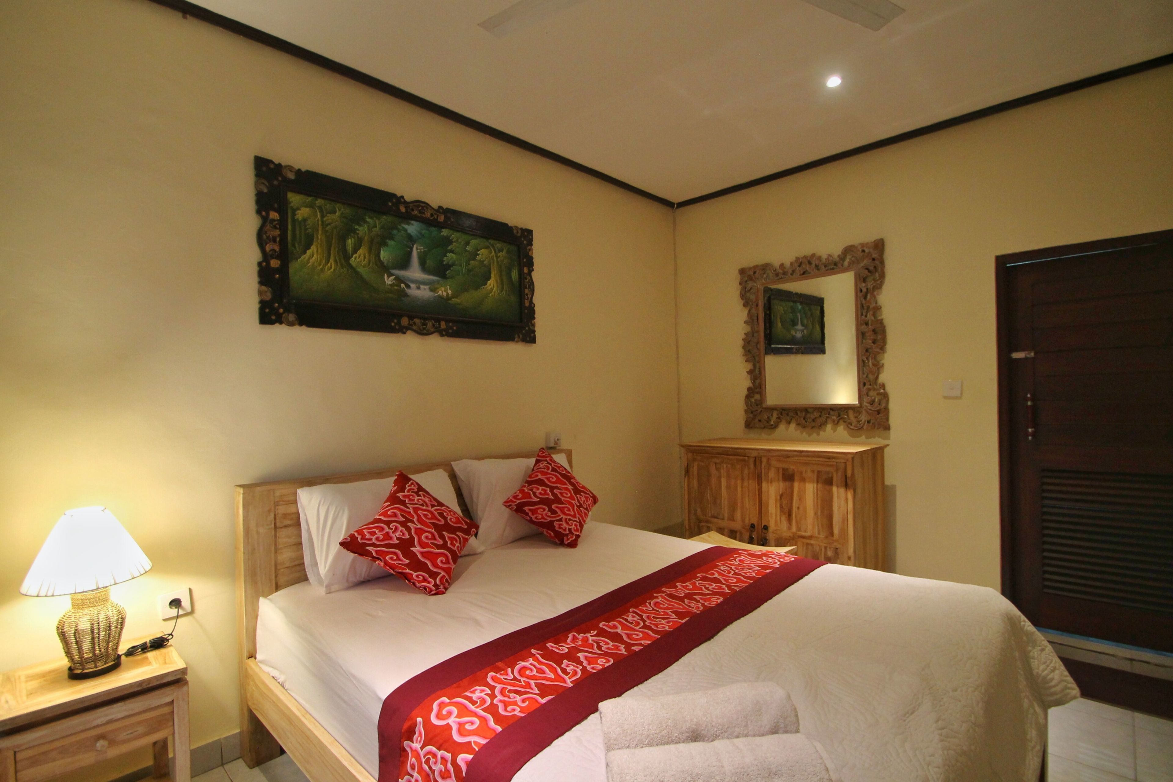 A neatly made bed with a red patterned bedspread and pillows, a bedside lamp, a framed painting, and a mirror on the wall.