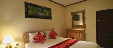 A neatly made bed with a red patterned bedspread and pillows, a bedside lamp, a framed painting, and a mirror on the wall.