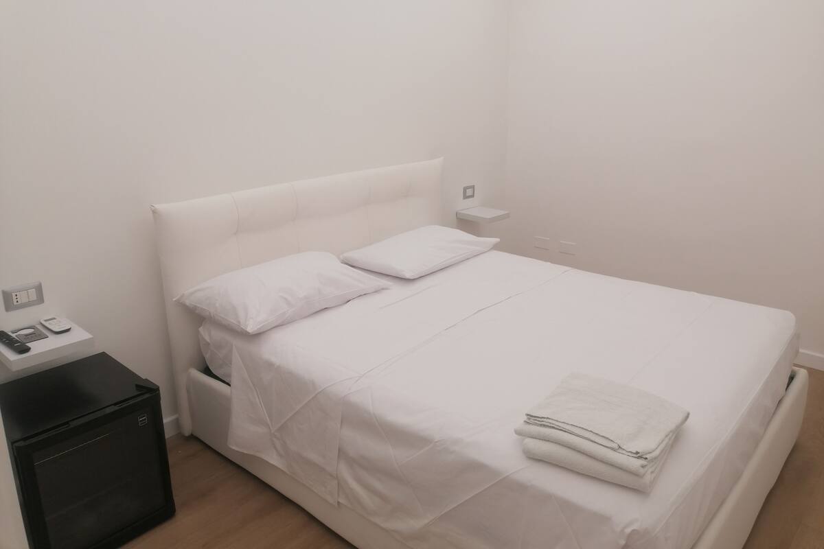 A neatly made bed with white linens and pillows, a small bedside table with a lamp, and a black storage unit.