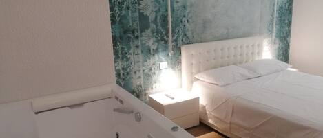 A bedroom with a white bed, a nightstand, and a white bathtub.