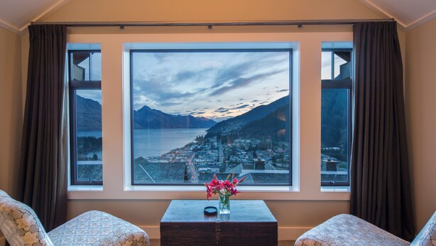 A room with two patterned armchairs, a wooden coffee table with a vase of flowers, and a large window offering a view of a lake and mountains.