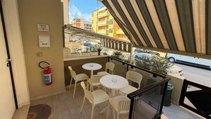 A balcony with white plastic chairs and tables, a fire extinguisher, a key box, and a view of the street and buildings. - The Smerald (Alghero)