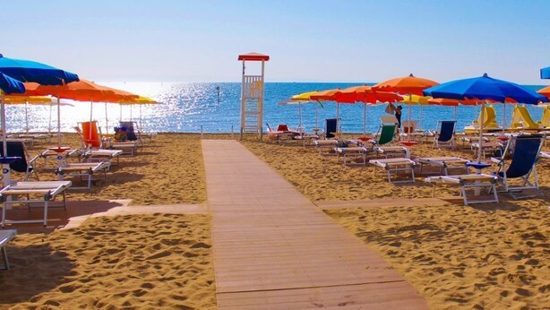 A beach with rows of sun loungers and umbrellas, a wooden walkway, and a lifeguard tower. - Appartamenti dei Fiori (Lignano Sabbiadoro)