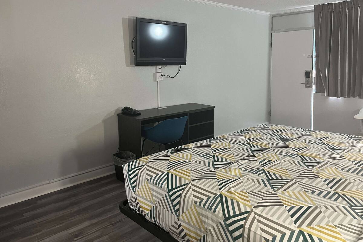 A hotel room with a bed, a desk, a chair, a television mounted on the wall, and a door with curtains.