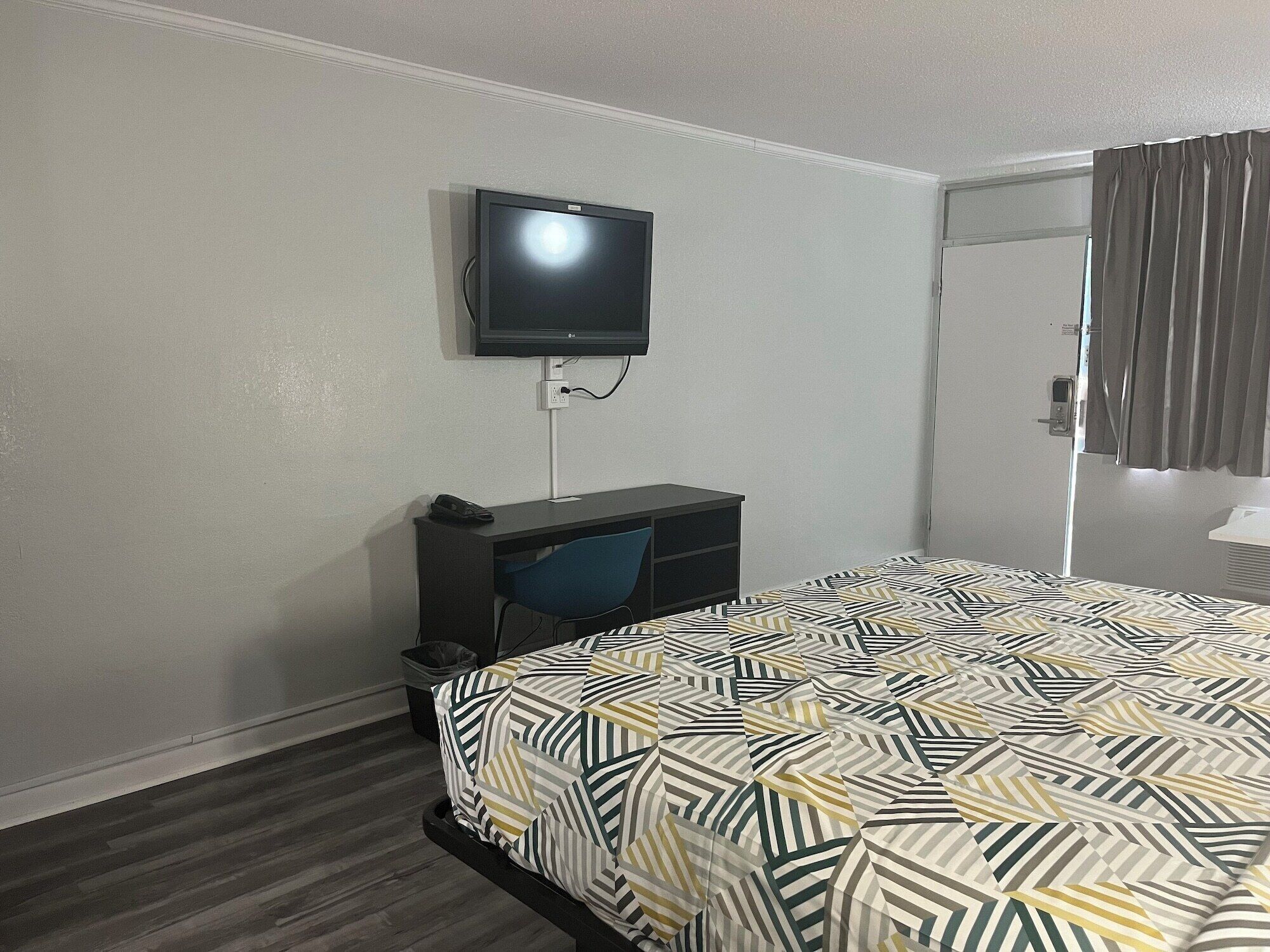 Habitación de hotel con cama, escritorio, silla, televisor montado en la pared y una puerta con cortinas.