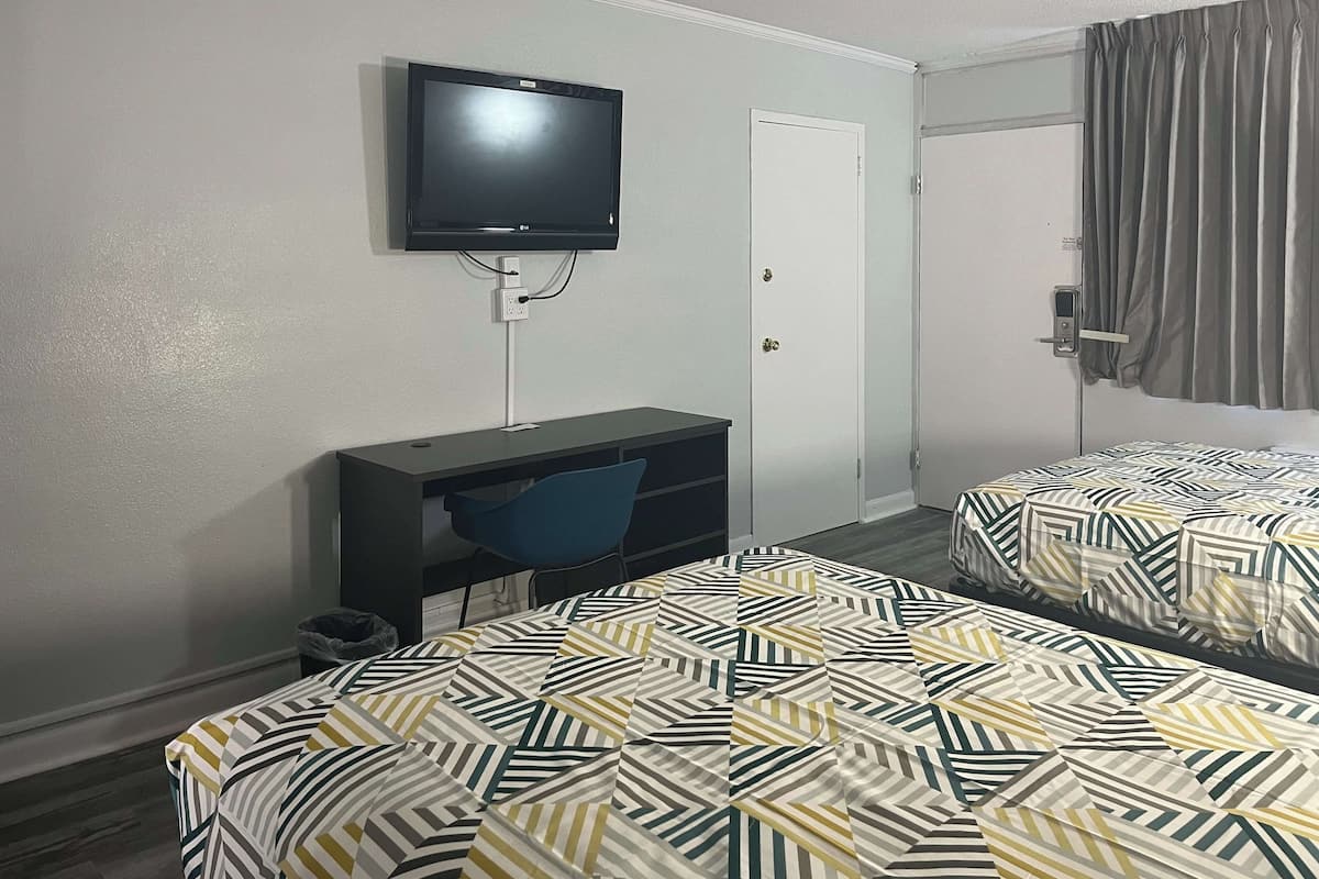 A hotel room with a TV mounted on the wall, a desk with a chair, and two beds with patterned bedding.
