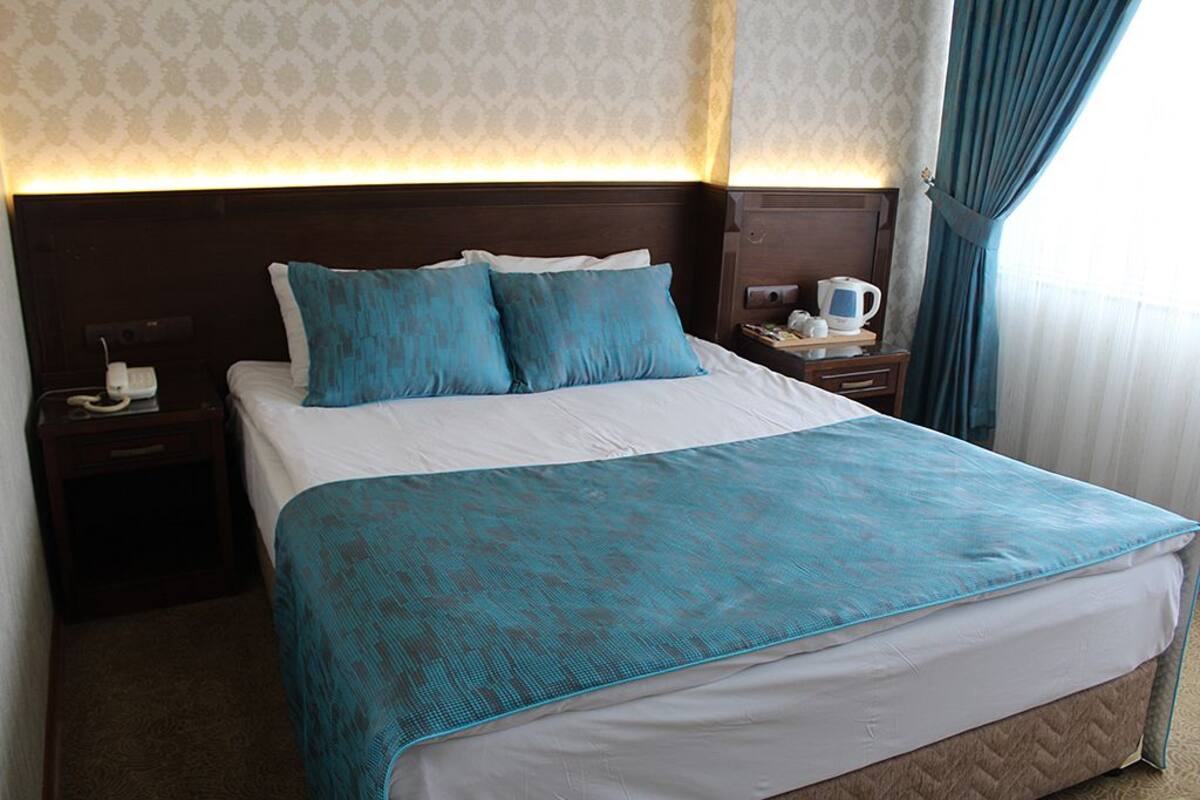 Habitación de hotel con una cama grande, una mesita de noche con teléfono y cafetera, y un ventanal con cortinas.