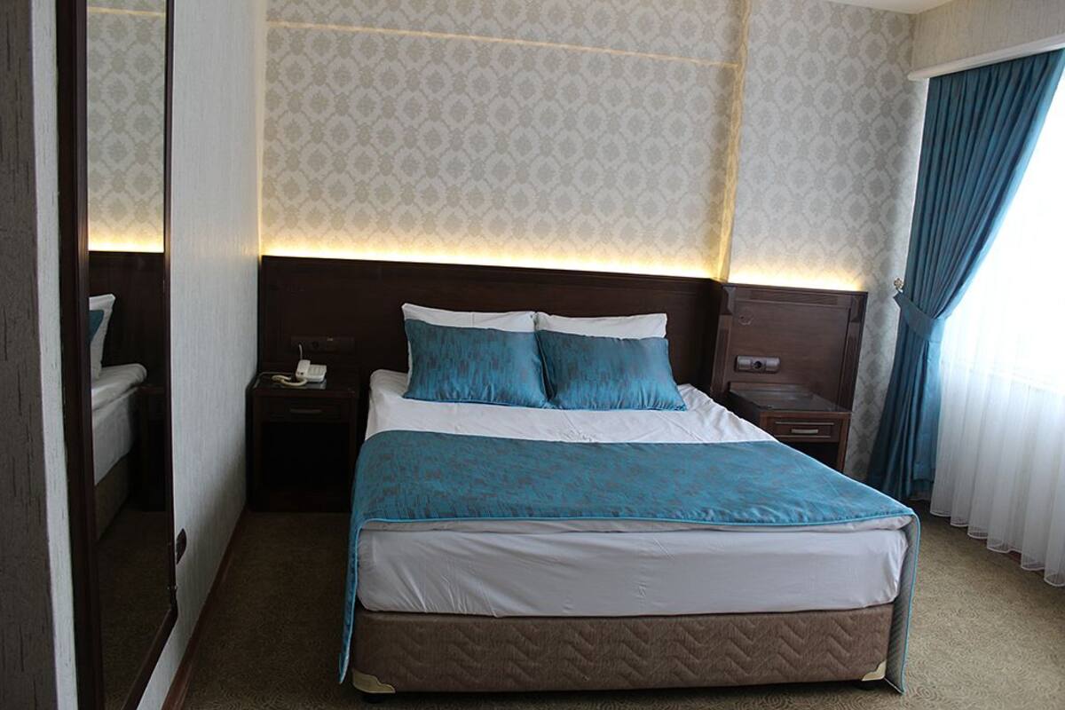 Una habitación de hotel con una cama grande, una mesita de noche con lámpara, y un ventanal con cortinas.
