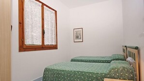 Una habitación pequeña y limpia con dos camas individuales, un escritorio de madera, una lámpara, un cuadro enmarcado en la pared y una ventana con cortinas de encaje.