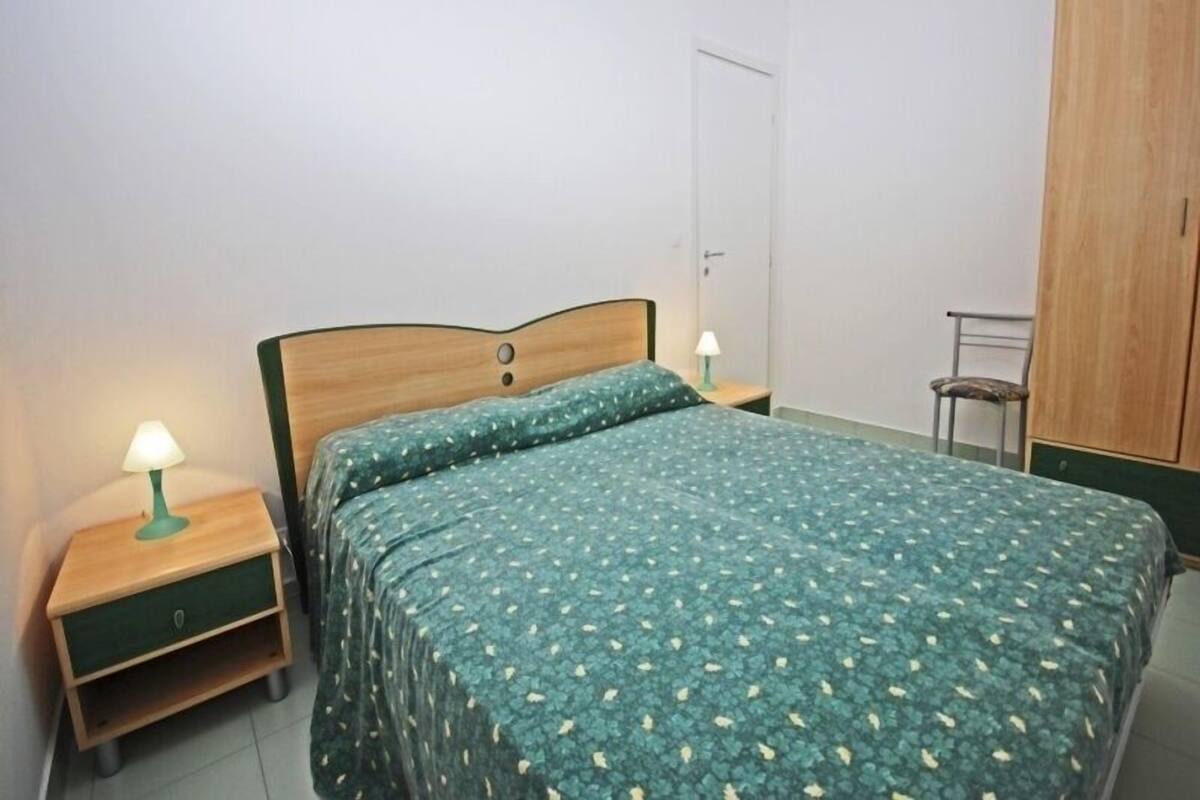 Een enkel bed met een beddegoed met een groen patroon, een houten hoofdbord, een nachtkastje met een groene lamp en een houten garderobe.