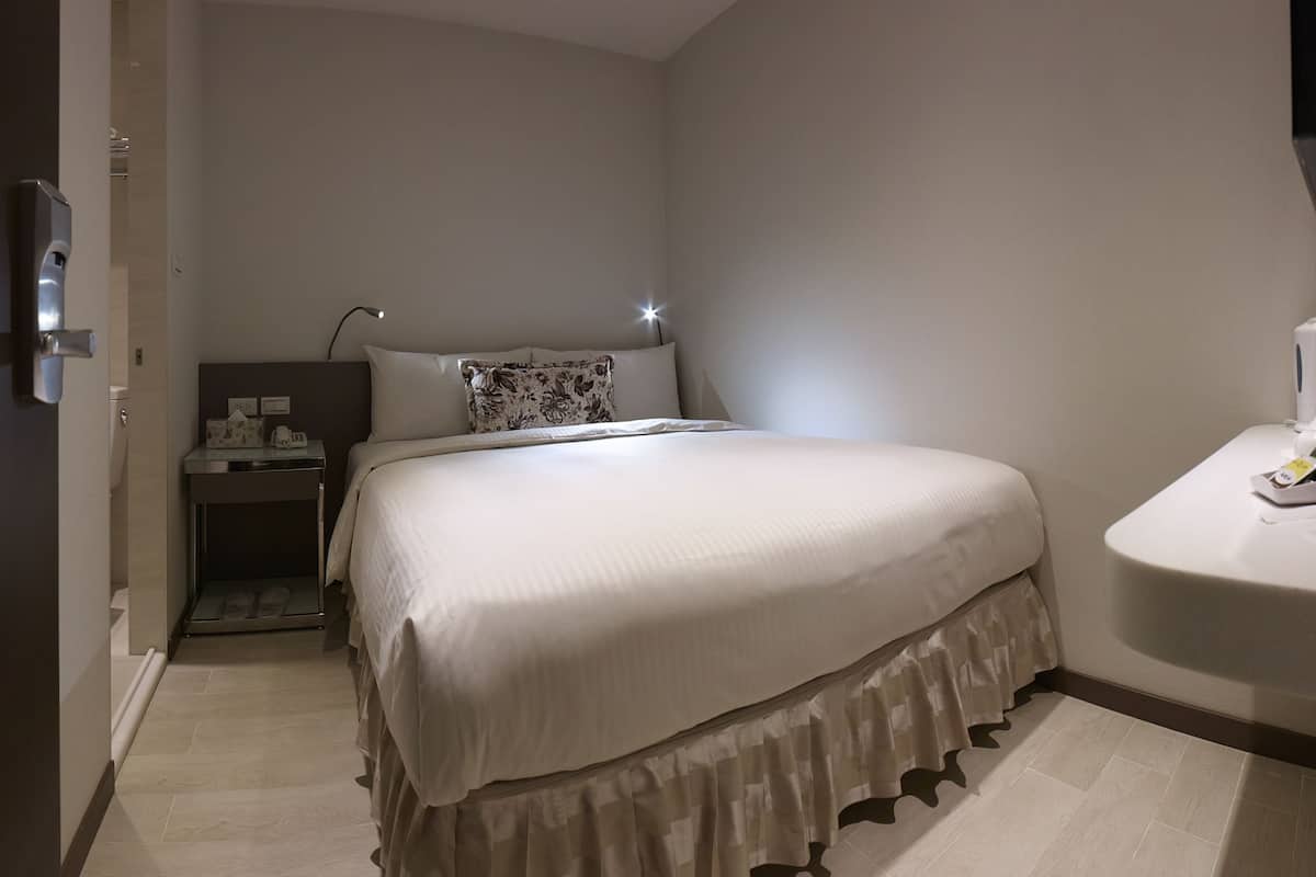 A hotel room with a bed, bedside table, lamp, and a small table with a phone and a bottle.