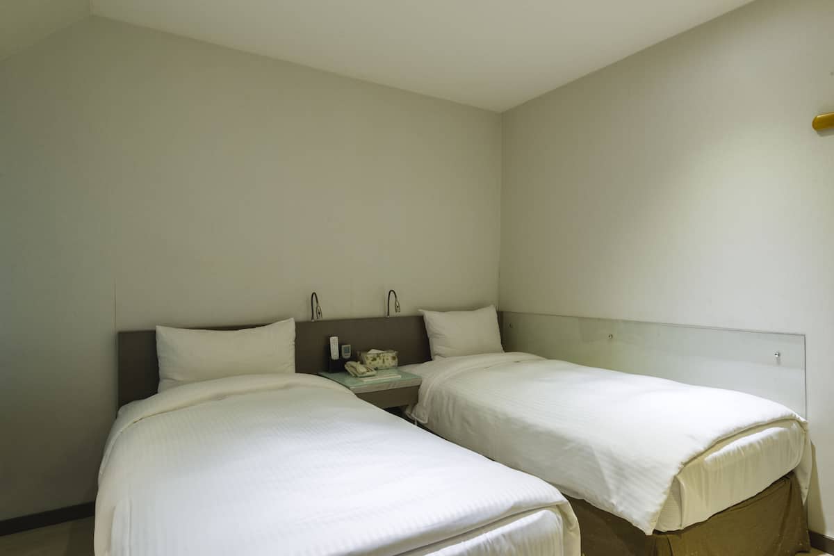 A hotel room with two single beds, a small bedside table with a telephone, and a plain wall.