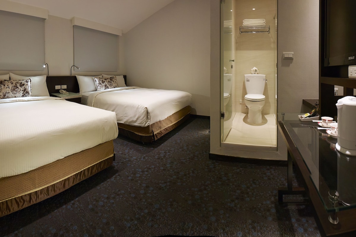 A hotel room with two beds, a glass-enclosed shower, a bedside table with a lamp, and a TV on a stand.
