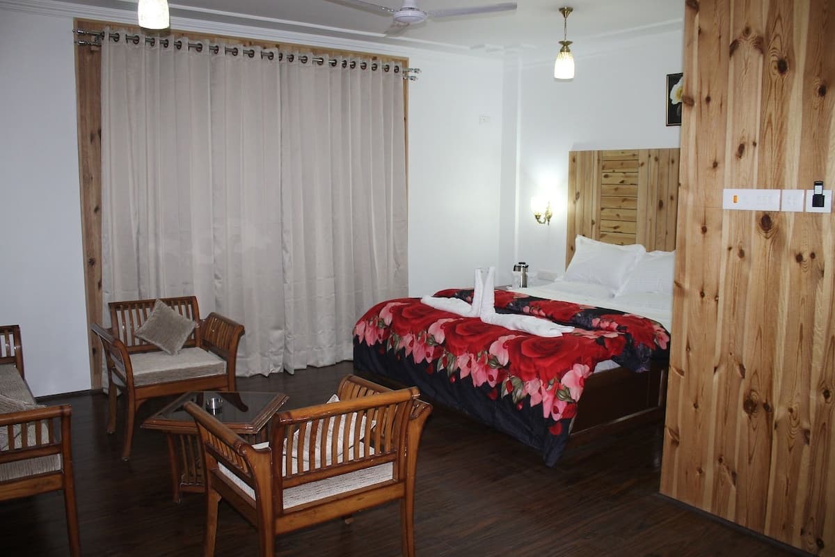 Quarto de hotel com cama, duas cadeiras de madeira, um guarda-roupa de madeira, um ventilador de teto e uma janela com cortinas.