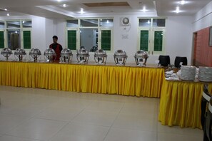 Banquet hall with a long table and silver service equipment, surrounded by windows and ceiling lights.