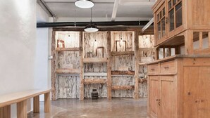 Una habitación amplia con techo alto, vigas de madera a la vista y suelo de concreto pulido. Hay bancos de madera a lo largo de una pared y un mueble de madera grande con puertas de cristal.