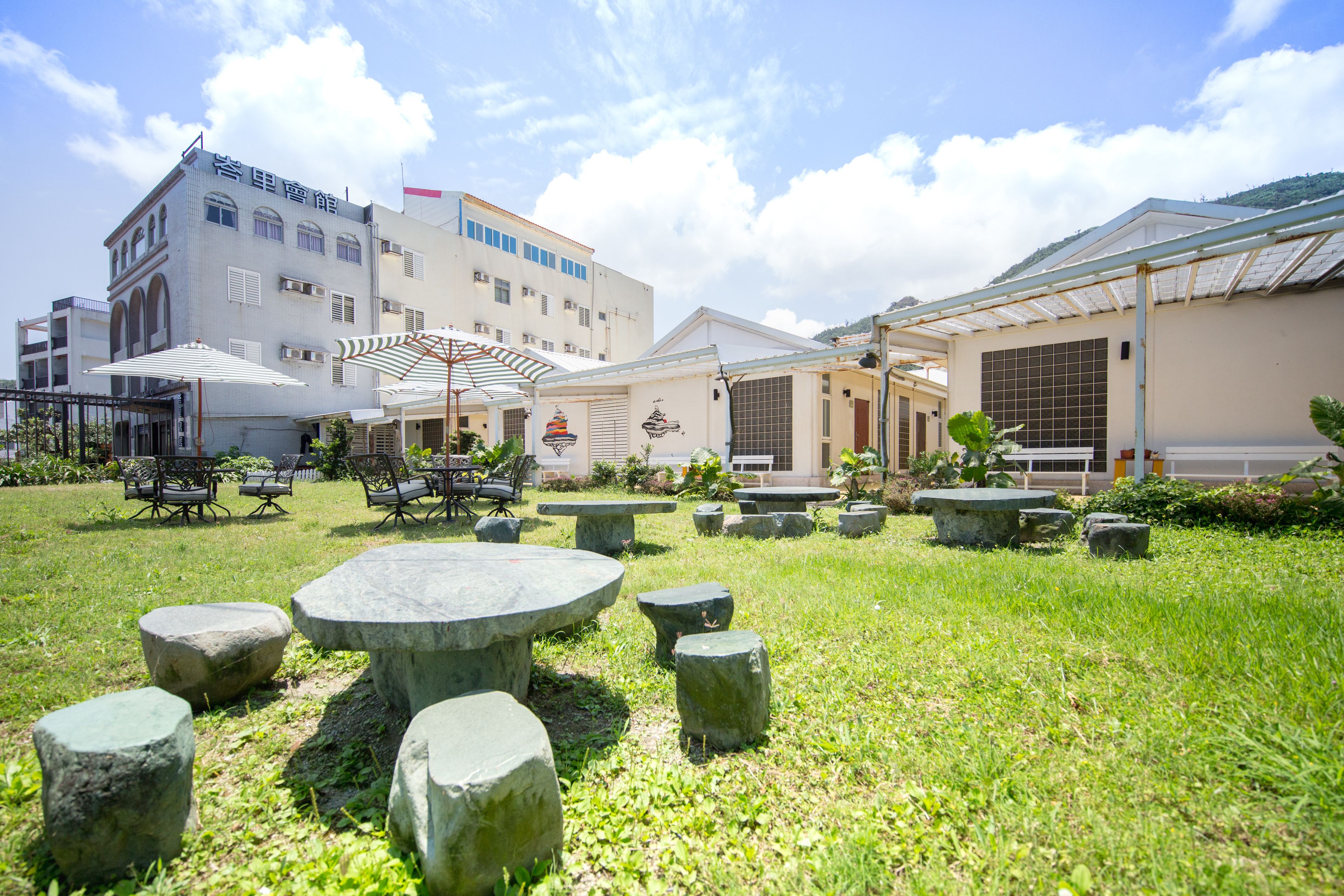 Pátio com mesas e cadeiras de pedra, rodeado por edifícios com grandes janelas e uma área coberta com guarda-sóis.