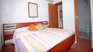 Quarto com cama de madeira, roupa de cama listrada e colorida, um quadro na parede e área de chuveiro visível através de uma porta aberta.