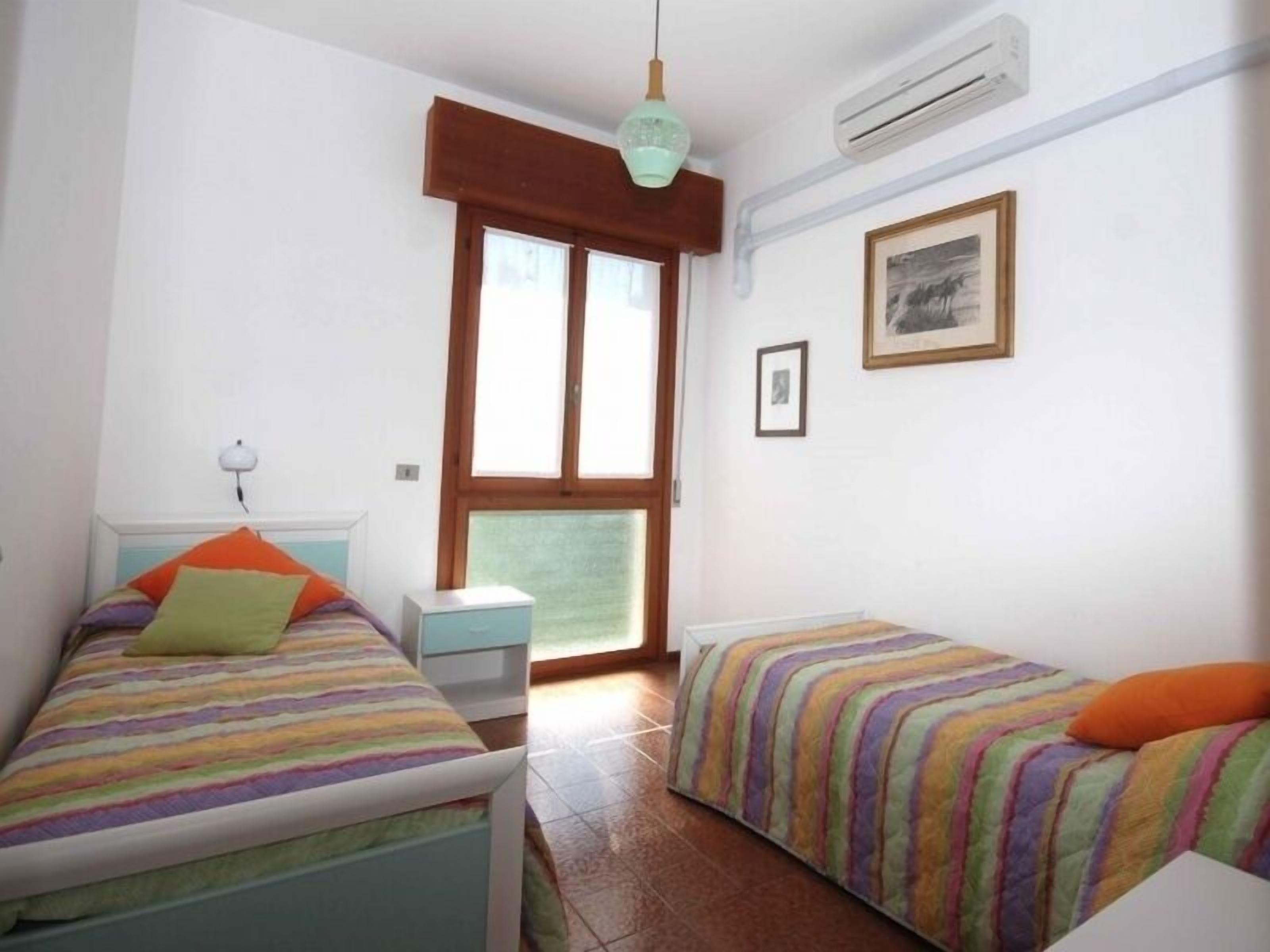 A small, well-lit room with two beds, a window, an air conditioner, and a framed picture on the wall.