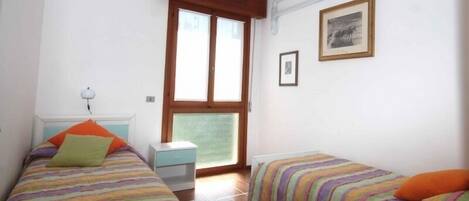 A small, well-lit room with two beds, a window, an air conditioner, and a framed picture on the wall.