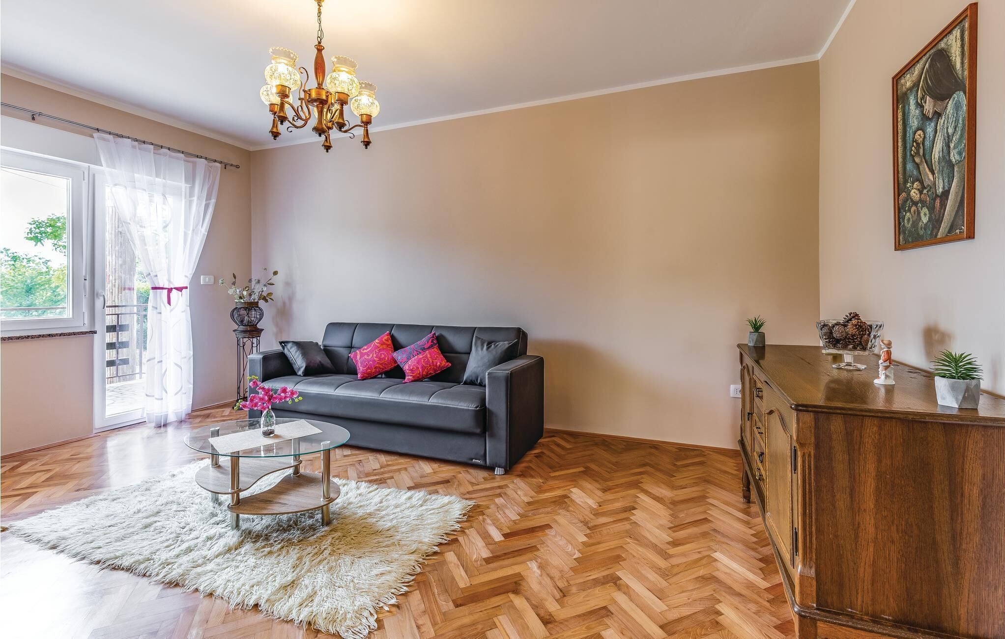 A living room with a black sofa, a glass coffee table, a wooden herringbone floor, a chandelier, and a painting on the wall.