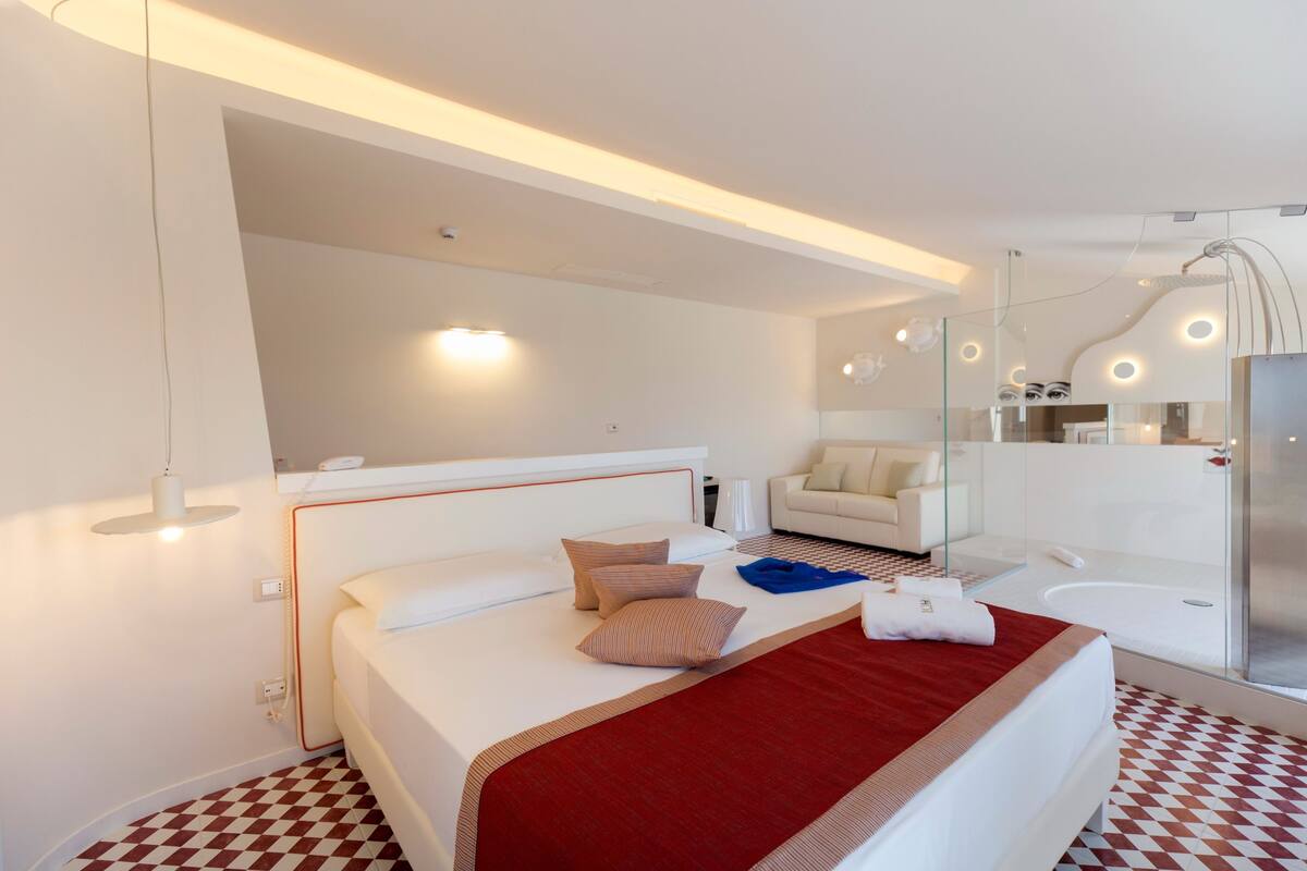 A modern hotel room with a large bed, a sofa, a bathroom with a glass shower, and a well-lit ceiling.