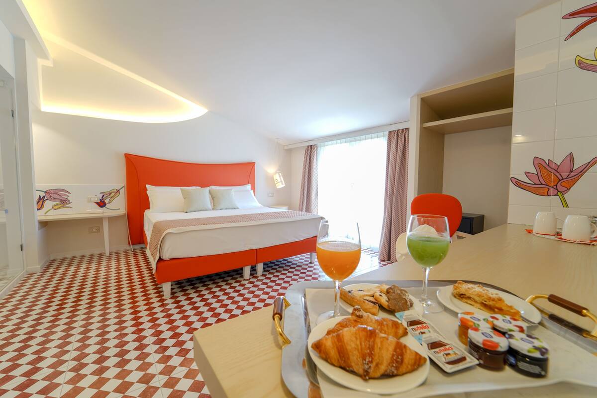 A hotel room with a checkered floor, a bed with an orange headboard, a dining table with breakfast items, and a window with curtains.