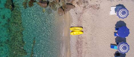 Vista aérea de una playa con agua turquesa cristalina, costa rocosa y varios sombrillas y sillas de playa.
