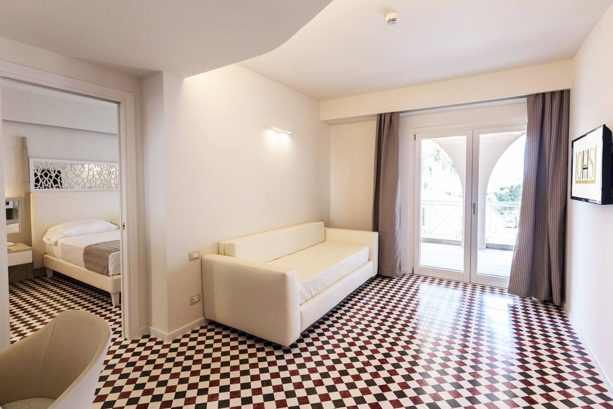 A hotel room with a checkered floor, a sofa, a bed, and a sliding glass door to the outside.