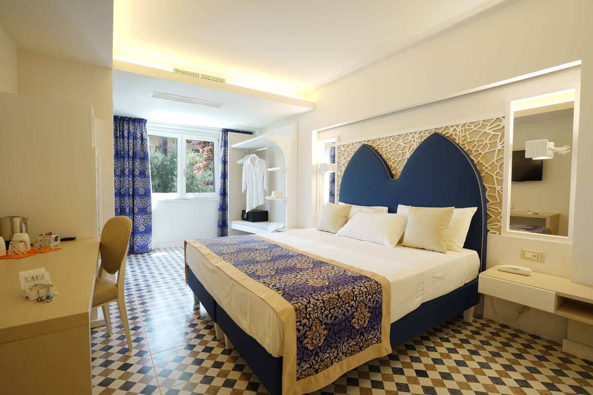A hotel room with a checkered floor, a bed with a patterned bedspread, a desk with a chair, a mirror, and a window with curtains.